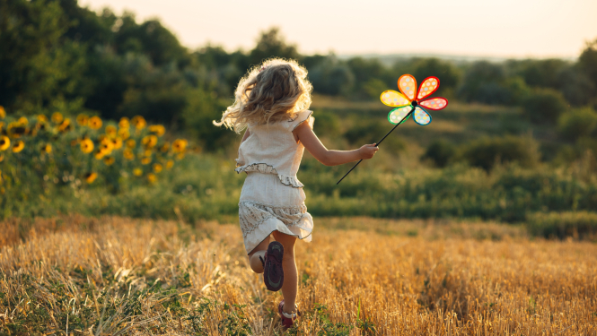 Bērns skrien pļavā, rokā krāsainas vējdzirnavas puķes formā