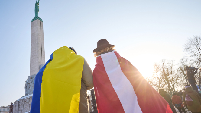 Pie Brīvības pieminekļa stāv divi cilvēki, viens uz pleciem tur Ukrainas karogu, otrs Latvijas karogu