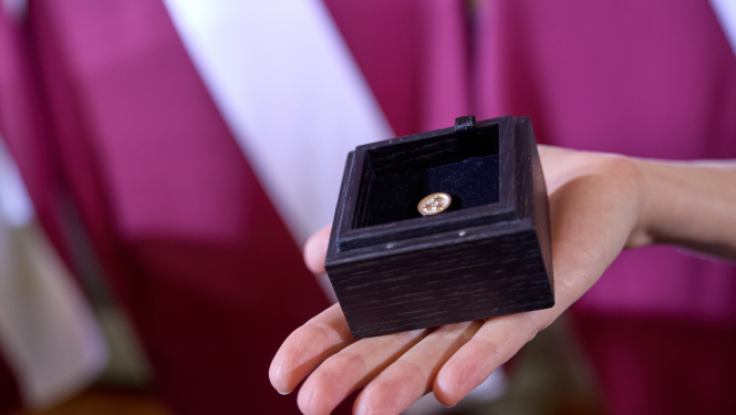 Uz Latvijas karoga fona plaukstā novietota koka kastīte, kurā ievietota Ministru kabineta balvas krūšu nozīmīte