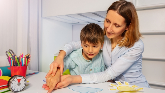 Mamma ar dēlu, kuram ir autisms. Uz galda krāsainas rotaļas. Mamma tur dēla roku 