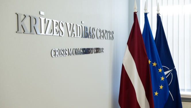Uz sienas ar sudraba burtiem uzraksts "Krīzes vadības centrs" tam līdzās Latvijas, Eiropas Savienības, NATO karogi