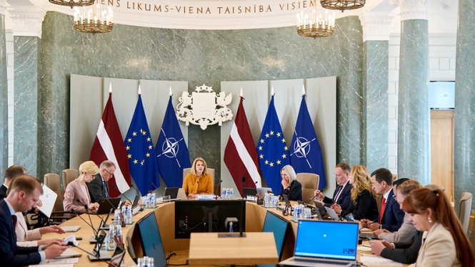 Ministru kabineta sēde. Ministru prezidente un ministri sēž pie sēžu galda. Fonā Latvijas, ES un NATO karogi un ģerbonis. 