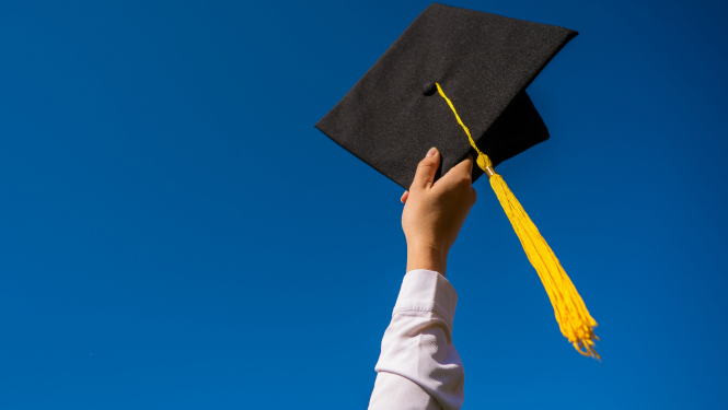 Rokā pacelta studenta izlaiduma cepure ar dzeltenu pušķi uz zila fona