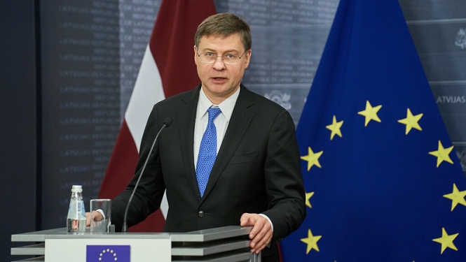 Eiropas Komisijas priekšsēdētājas izpildvietnieks Valdis Dombrovskis preses konferenču telpā. Fonā Latvijas un Eiropas Savienības karogi. 