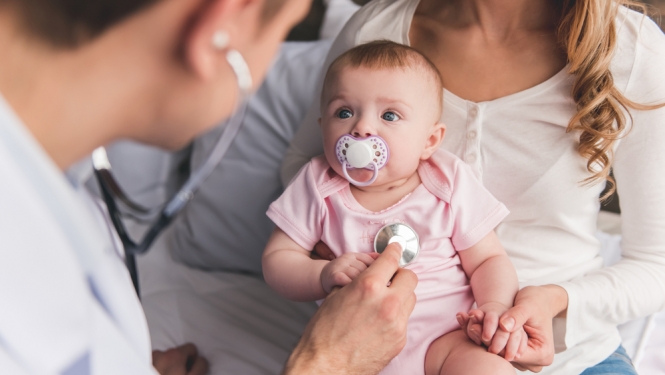 Ģimenes ārsts pārbauda mazuli ar stetoskopu. Mazulis ir rokās mammai. 