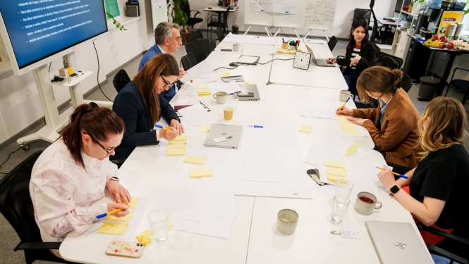 Cilvēki piedalās inovācijas sprintā, sēž ap galdu, sarunājas, raksta