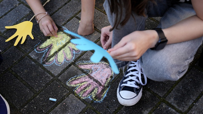 Bērni, kas ar krītiņiem uz bruģa zīmē rociņas krāsainas