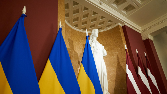 Pa labi trīs Ukrainas karogi, tālāk, statuja Taisnība un trīs Latvijas karogi.