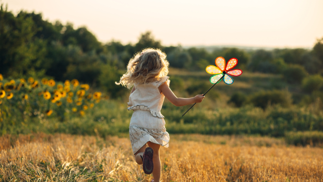 Bērns skrien pļavā, rokā krāsainas vējdzirnavas puķes formā
