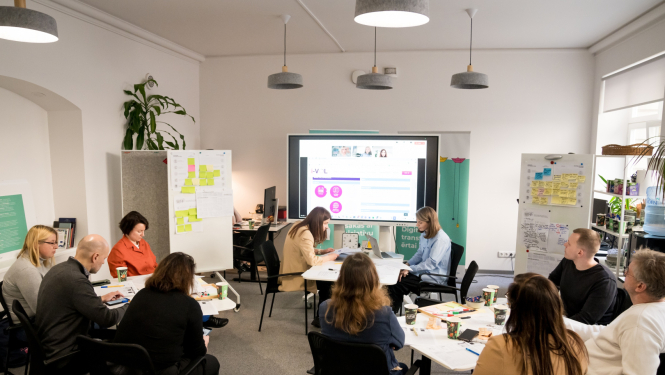 Inovāciju sprints - dalībnieki sēž pie galdiem, sarunājas un vēro ekrānu
