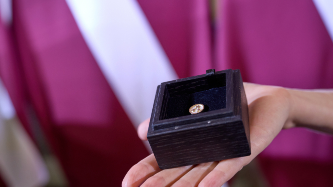 Uz Latvijas karoga fona plaukstā novietota koka kastīte, kurā ievietota Ministru kabineta balvas krūšu nozīmīte