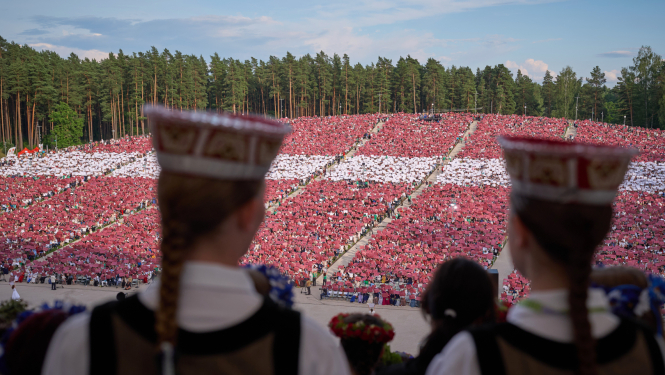 Priekšplānā meiteņu silueti latviešu tautastērpos, tālāk skats uz Mežaparka estrādes skatītāju zonu, kurā skatītāji izveidojuši Latvijas karogu, paceļot gaisā krāsainas papīra lapas