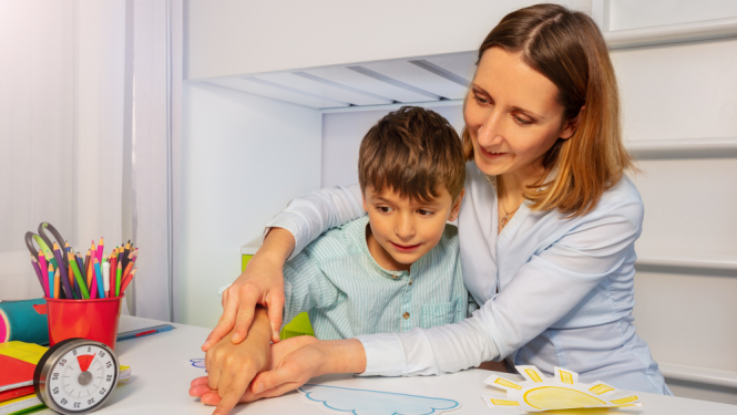 Mamma ar dēlu, kuram ir autisms. Uz galda krāsainas rotaļas. Mamma tur dēla roku 
