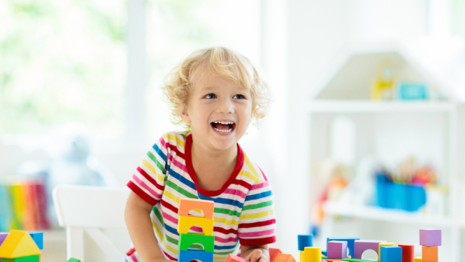 Bērns rotaļās ar krāsainiem klucīšiem un smaida. Gaišs fons un fonā plaukts ar klucīšiem. 