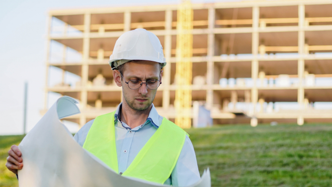 Būvnieks ar ķiveri pēta dokumentāciju. Fonā koka konstrukcija. 