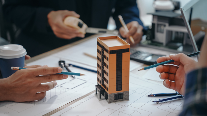 Daudzdzīvokļu nama modelis uz galda, tam apkārt rasējumi un cilvēku rokas, kuras tur zīmuļus