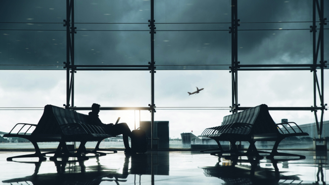 Cilvēks sēž lidostas uzgaidāmajā zālē pie lieliem logiem, strādājot ar portatīvo datoru. Tuvumā atrodas ceļojuma soma. Logā redzama lidmašīna, kas paceļas un debesīs pelēki mākoņi.
