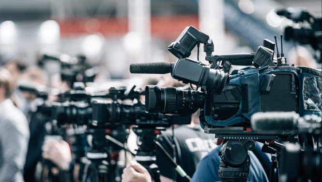 Attēlā redzamas vairākas profesionālas videokameras, kas novietotas uz statīviem un fokusētas uz notikumu vai preses konferenci. Dažas kameras ir aprīkotas ar mikrofonu un citām papildierīcēm.