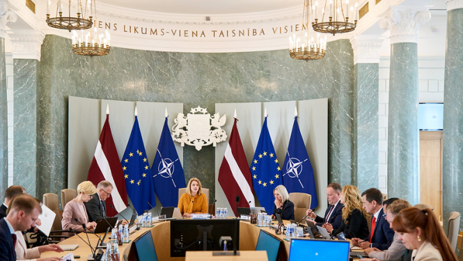 Ministru kabineta sēde. Ministru prezidente un ministri sēž pie sēžu galda. Fonā Latvijas, ES un NATO karogi un ģerbonis. 