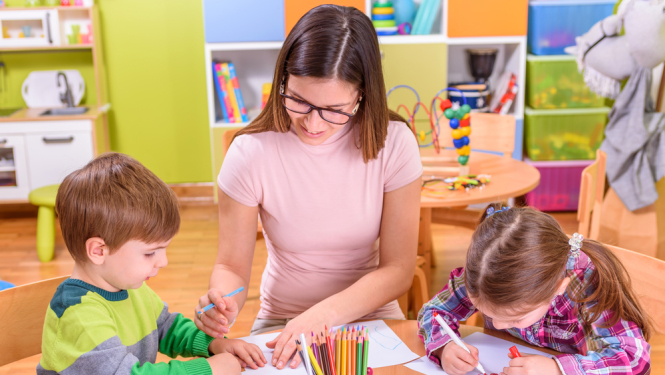 Pirmsskolas skolotāja un bērni pie galda. Skolotāja palīdz bērniem zīmēt.