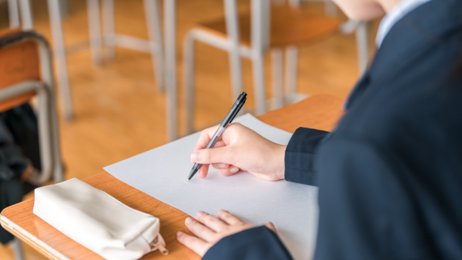 Skolēns rokā tur pildspalvu, uz rakstāmgalda balta lapa un rakstāmlietu maciņš