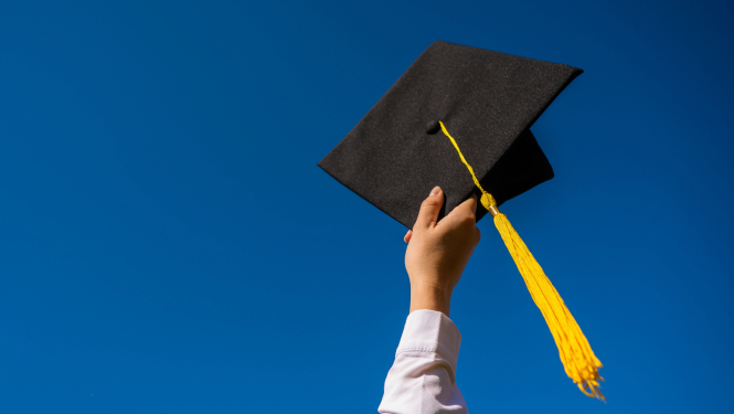 Rokā pacelta studenta izlaiduma cepure ar dzeltenu pušķi uz zila fona