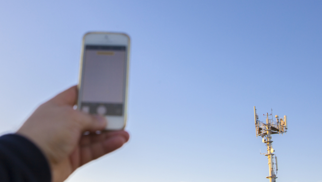 Cilvēks tur rokā mobilo telefonu, fonā sakaru tornis