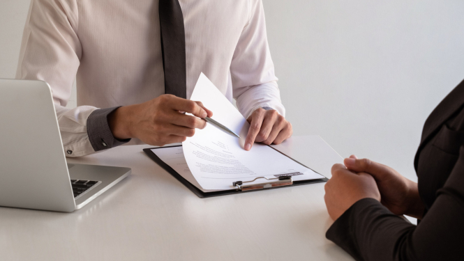 Foto: 2 cilvēki pie galda, viens tur rokā mapīti, uz galda dators