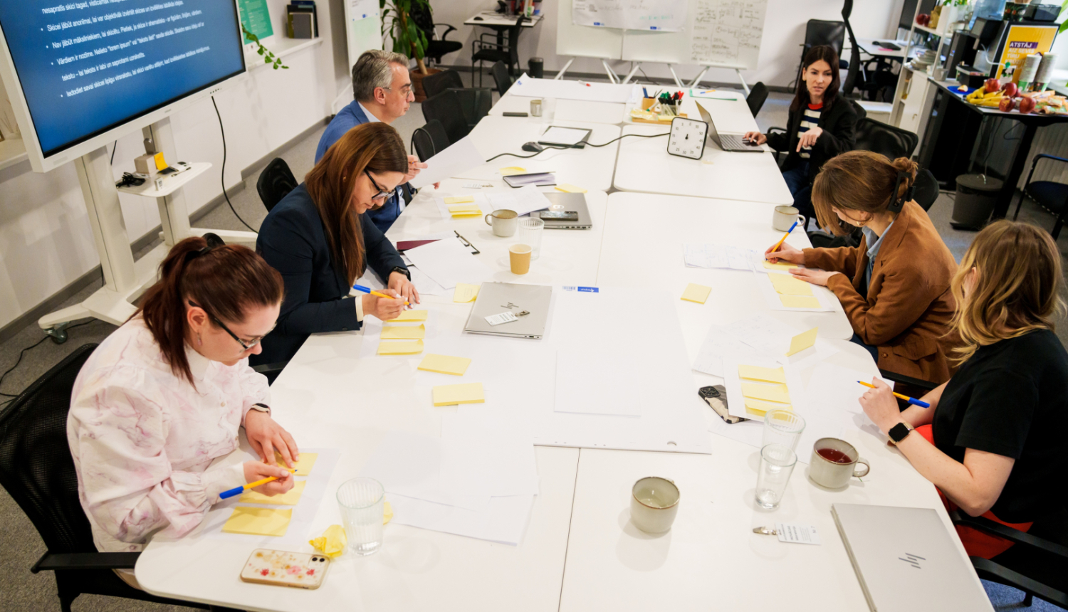 Cilvēki piedalās inovācijas sprintā, sēž ap galdu, sarunājas, raksta