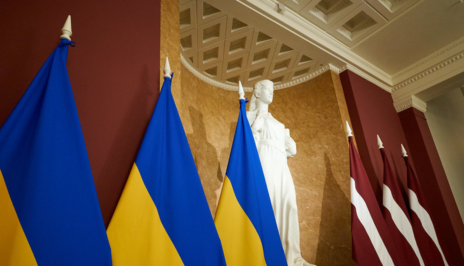 Pa labi trīs Ukrainas karogi, tālāk, statuja Taisnība un trīs Latvijas karogi.