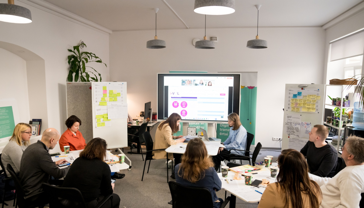 Inovāciju sprints - dalībnieki sēž pie galdiem, sarunājas un vēro ekrānu
