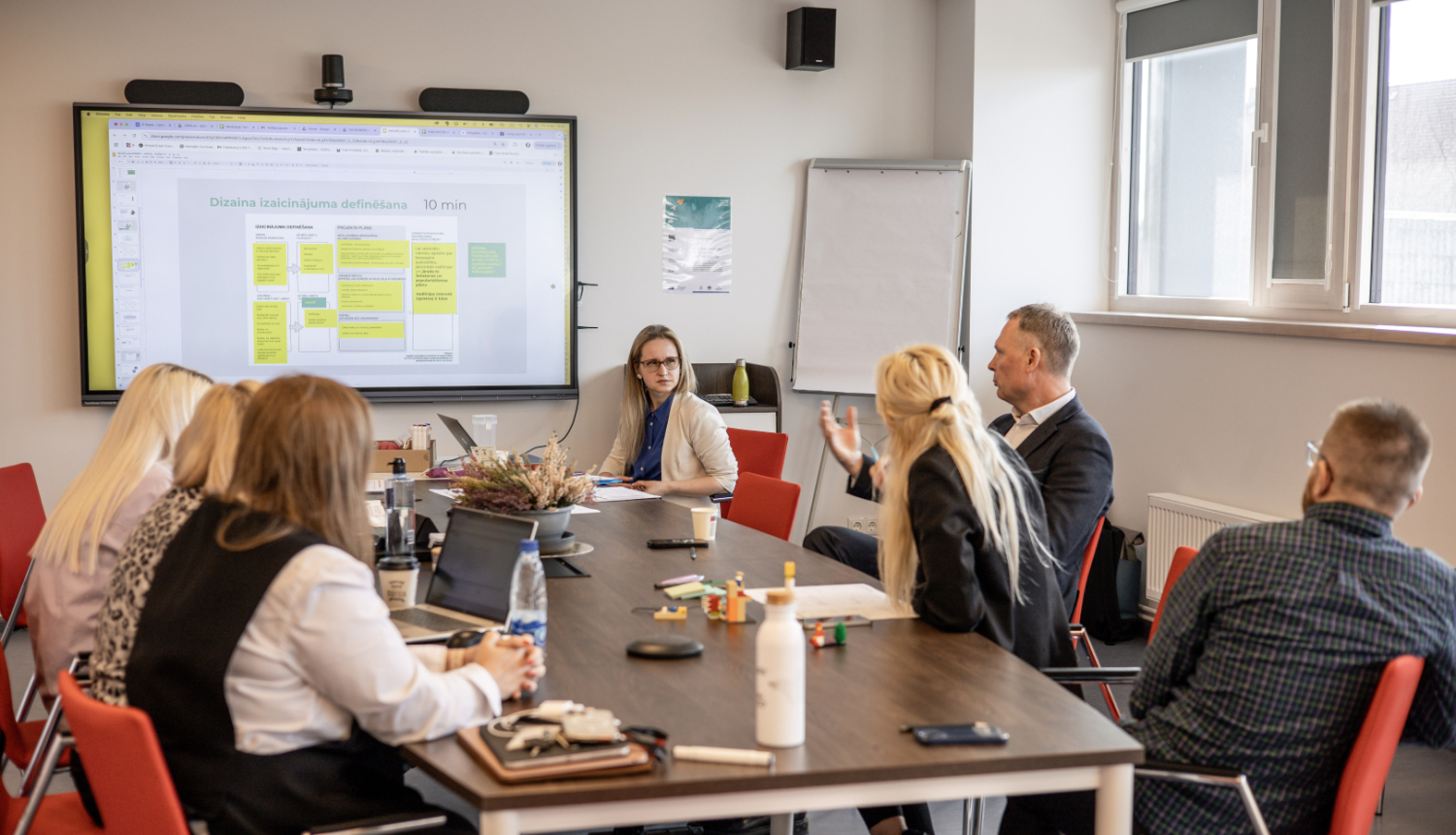 Inovāciju sprints - dalībnieki sēž pie galda, sarunājas un vēro ekrānu