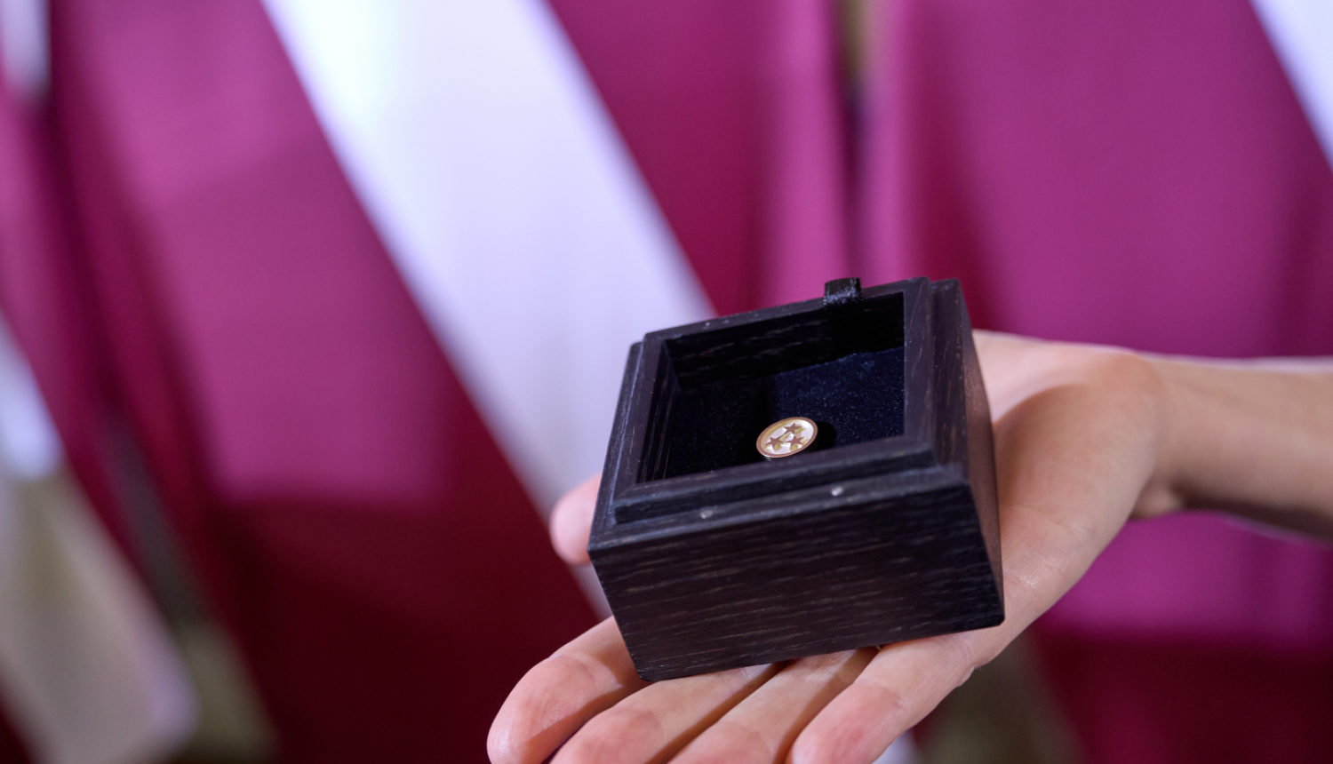 Uz Latvijas karoga fona plaukstā novietota koka kastīte, kurā ievietota Ministru kabineta balvas krūšu nozīmīte