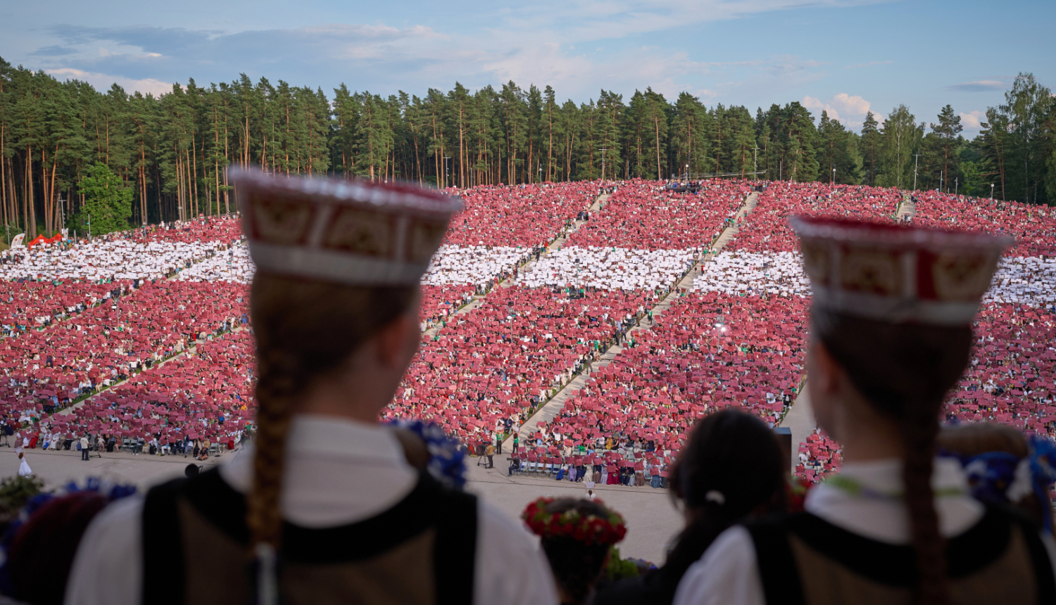 Priekšplānā meiteņu silueti latviešu tautastērpos, tālāk skats uz Mežaparka estrādes skatītāju zonu, kurā skatītāji izveidojuši Latvijas karogu, paceļot gaisā krāsainas papīra lapas