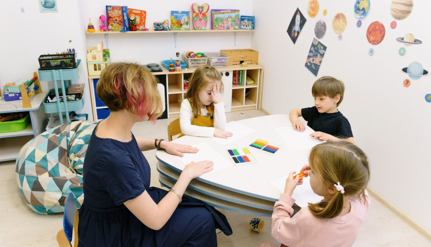 Mazi bērni ap galdu un sieviete. Fonā plaukti ar krāsainām mantiņām. 