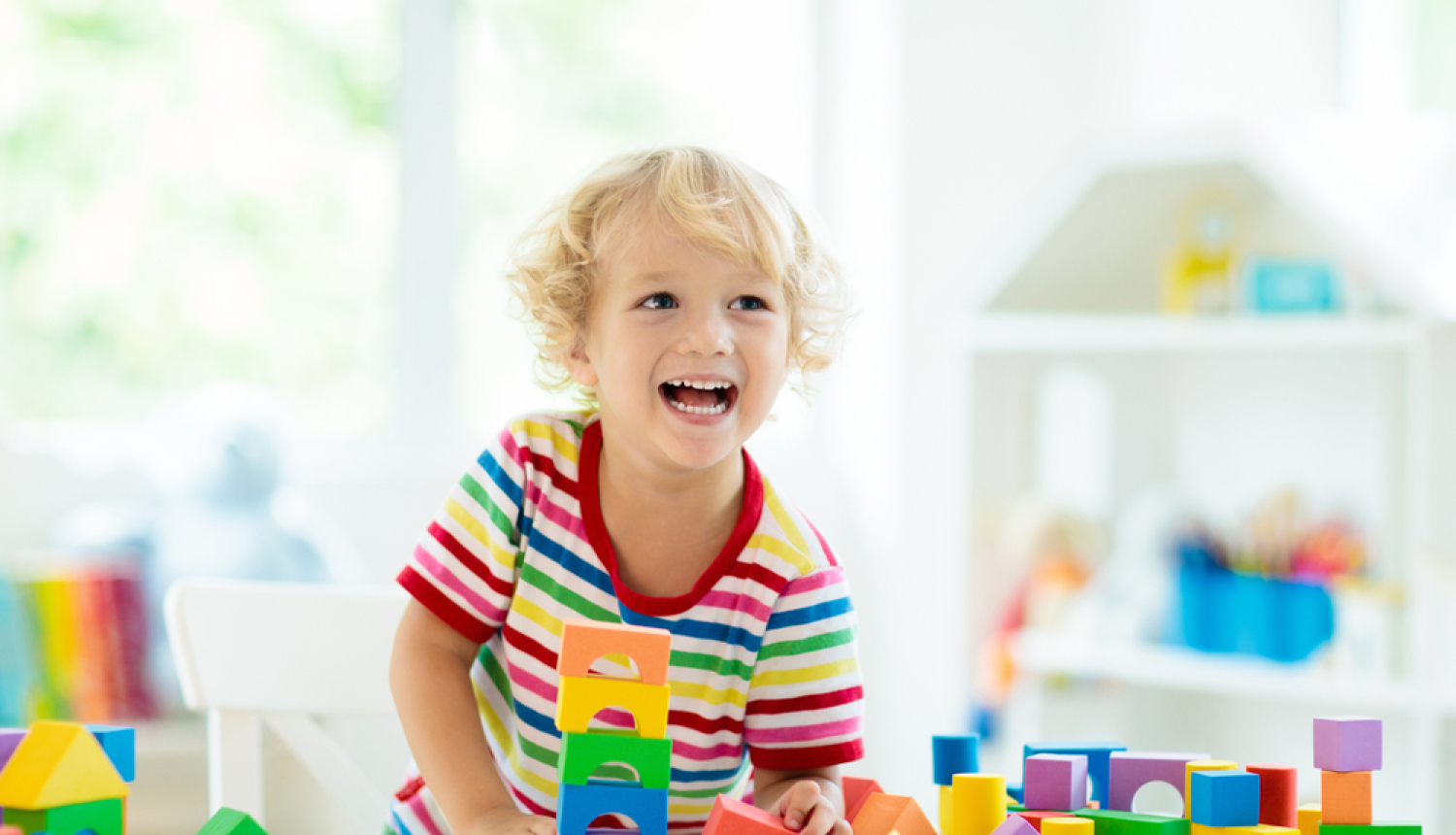 Bērns rotaļās ar krāsainiem klucīšiem un smaida. Gaišs fons un fonā plaukts ar klucīšiem. 