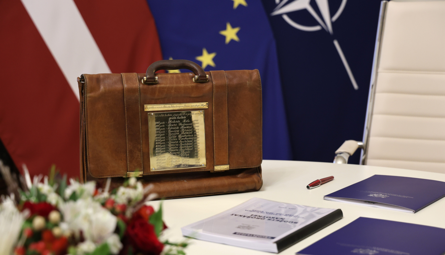 Uz galda novietots brūnais ādas portfelis, kurā tradicionāli uz Saeimu nogādā valsts budžeta likumprojektu nākamajam gada. Uz portfeļa metāla plāksne, uz kuras iegravēti finanšu ministru vārdi. Aiz portfeļa Latvijas, Eiropas Savienības un NATO karogi.