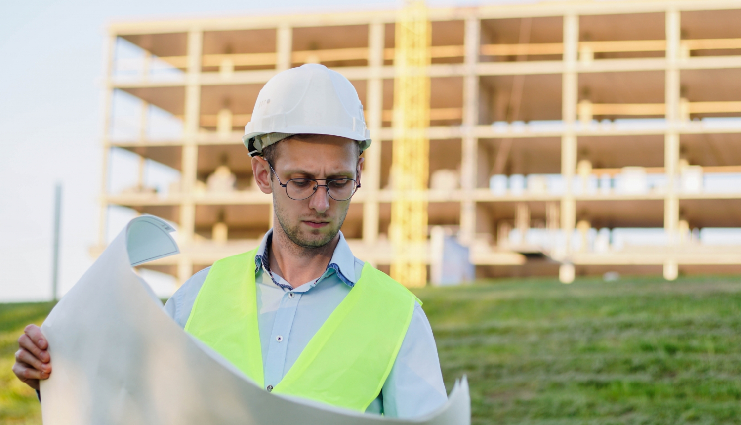 Būvnieks ar ķiveri pēta dokumentāciju. Fonā koka konstrukcija. 