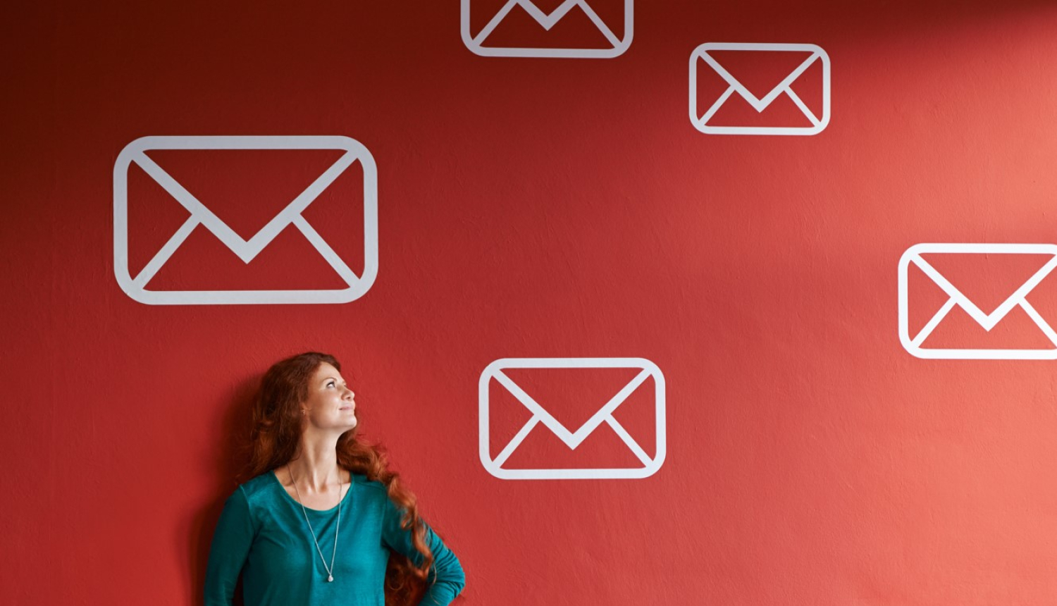 Sieviete ar gariem, sarkanīgiem matiem, ģērbusies zaļā blūzē, stāv pie sarkanas sienas ar uzzīmētām baltām vēstuļu ikonām, skatoties uz augšu.