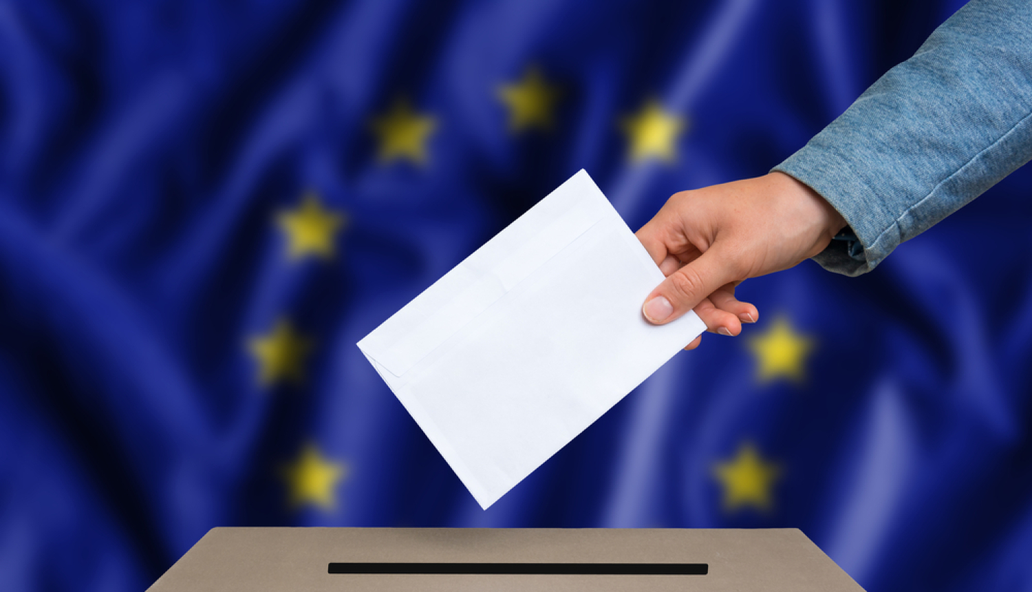 Eiropas Parlamenta vēlēšanas. Roka met kastē aploksni. Fonā Eiropas Savienības karogs. 