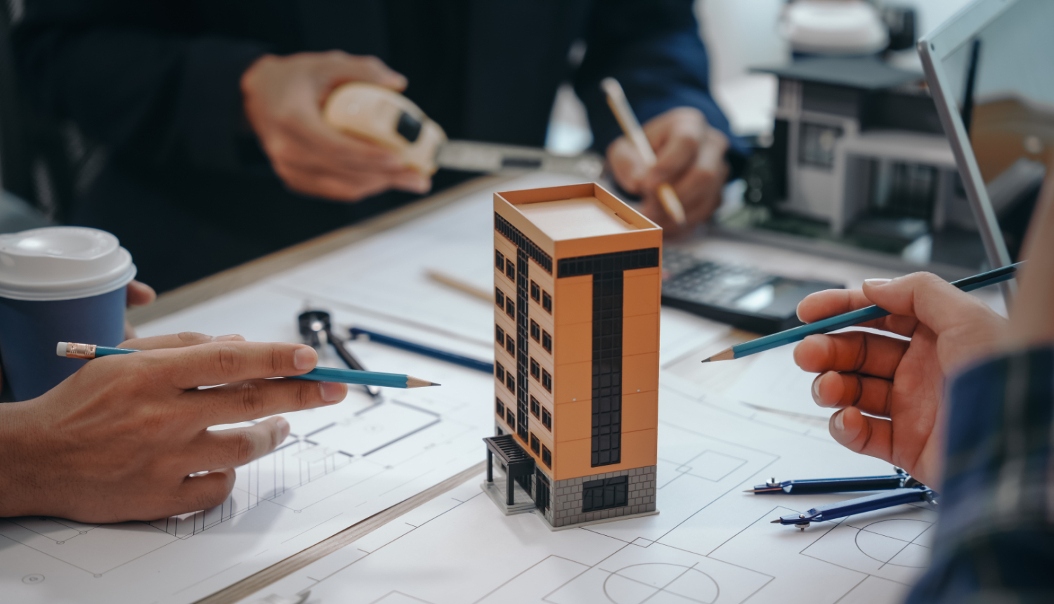 Daudzdzīvokļu nama modelis uz galda, tam apkārt rasējumi un cilvēku rokas, kuras tur zīmuļus