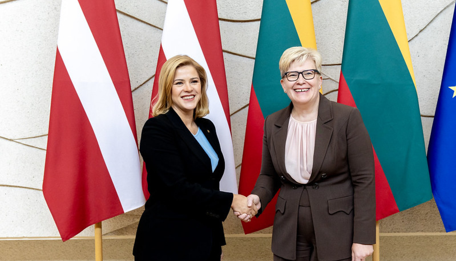 Evika Siliņa un Ingrīda Šimonīte sasveicinās. Fonā Latvijas un Lietuvas karogi. 