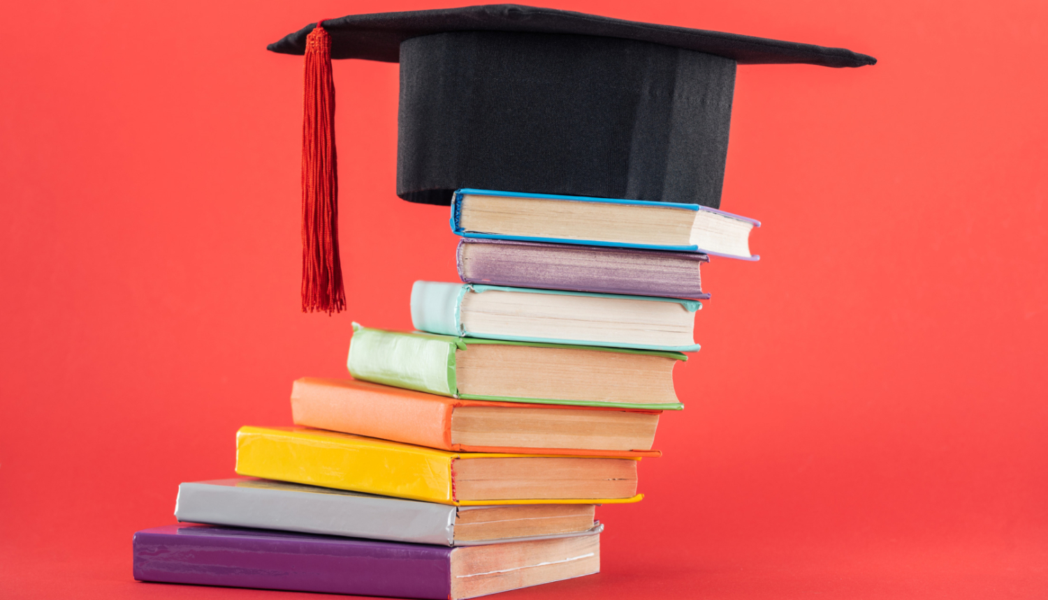 Uz krāsainu grāmatu kaudzītes novietota augstskolas absolventa cepure ar sarkanu pušķi