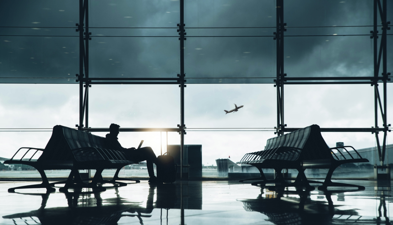 Cilvēks sēž lidostas uzgaidāmajā zālē pie lieliem logiem, strādājot ar portatīvo datoru. Tuvumā atrodas ceļojuma soma. Logā redzama lidmašīna, kas paceļas un debesīs pelēki mākoņi.