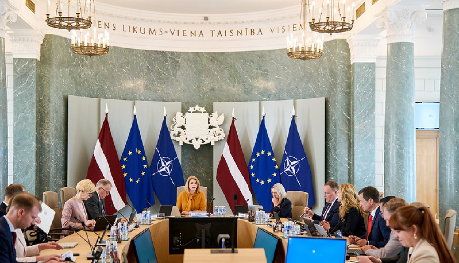 Ministru kabineta sēde. Ministru prezidente un ministri sēž pie sēžu galda. Fonā Latvijas, ES un NATO karogi un ģerbonis. 