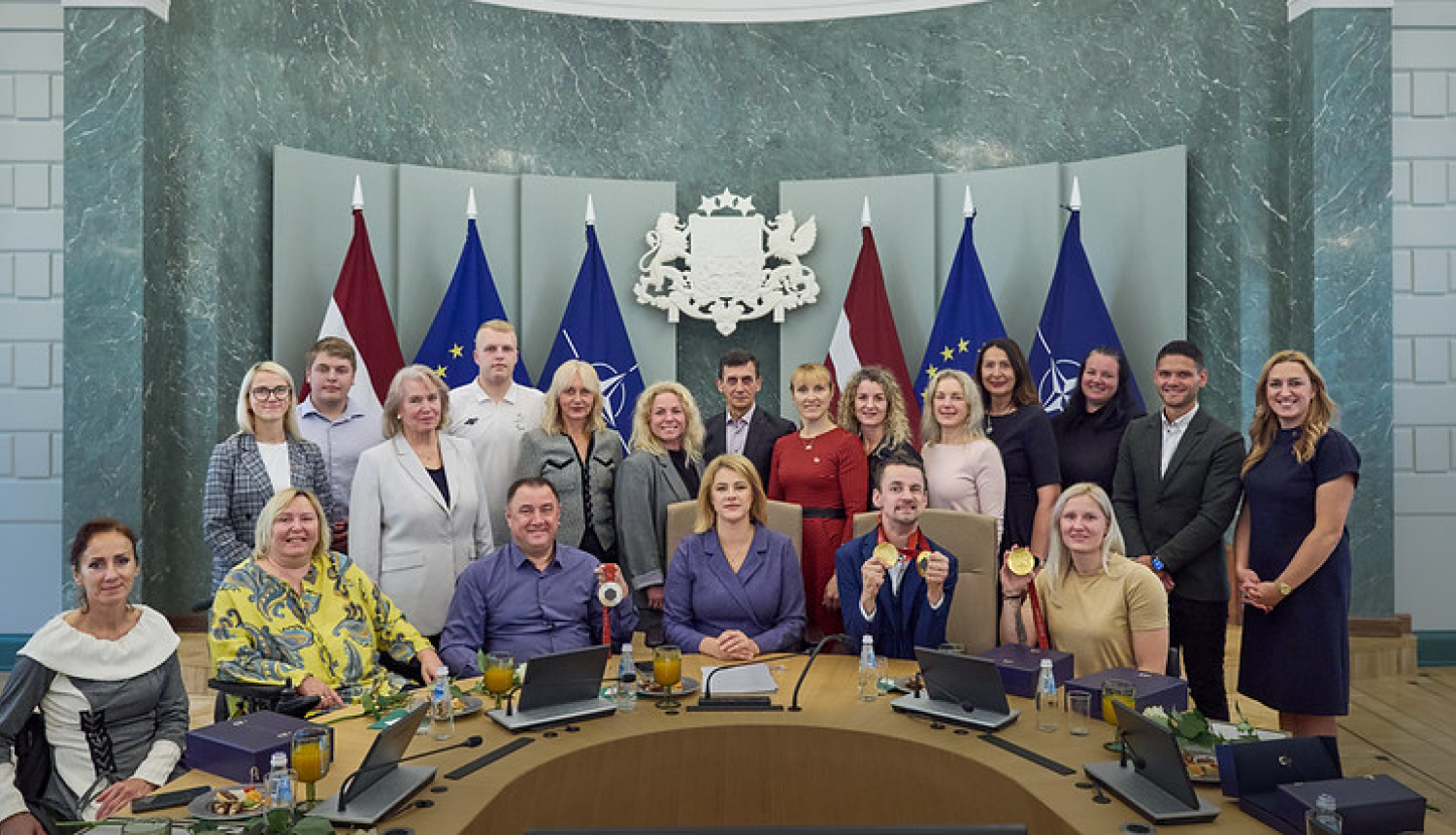 Kopbilde MK sēžu zālē, kurā redzami Latvijas paraolimpieši, treneri, delegācijas dalībnieki un centrā Ministru prezidente. 