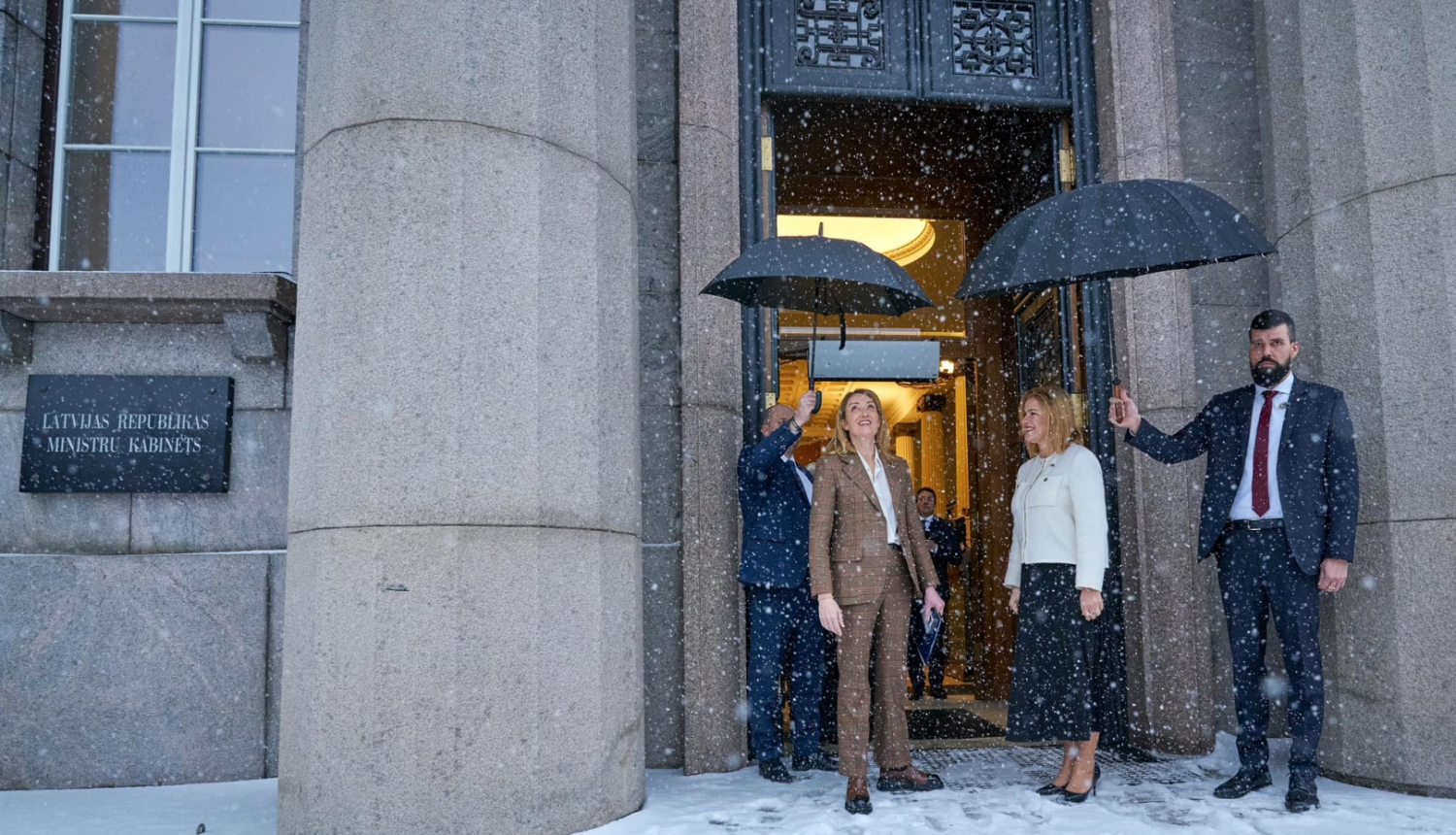 Roberta Metsola un Evika Siliņa pie valdības nama. Snieg sniegs un 2 vīrieši tur virs viņām lietussargus. 