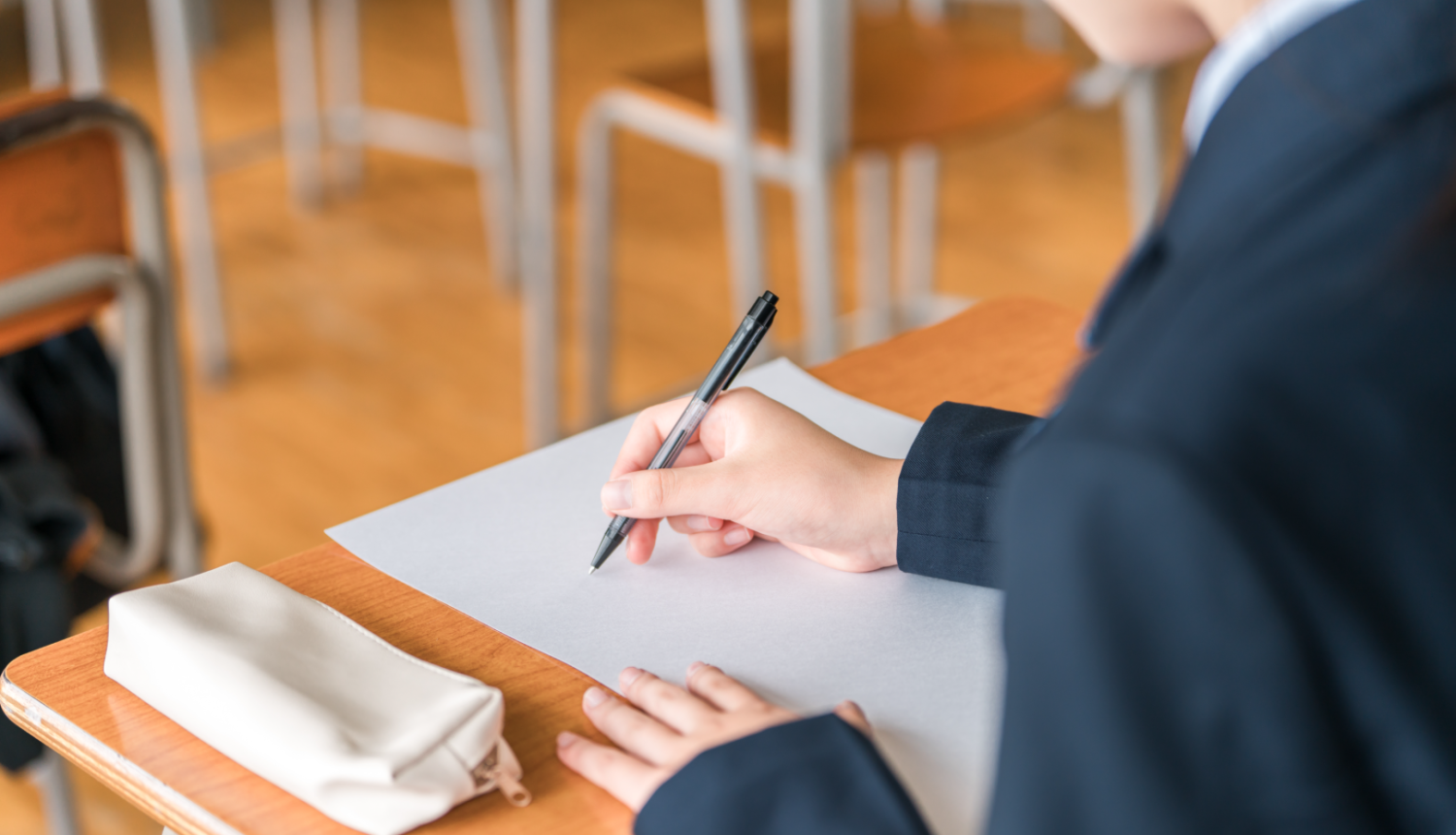 Skolēns rokā tur pildspalvu, uz rakstāmgalda balta lapa un rakstāmlietu maciņš