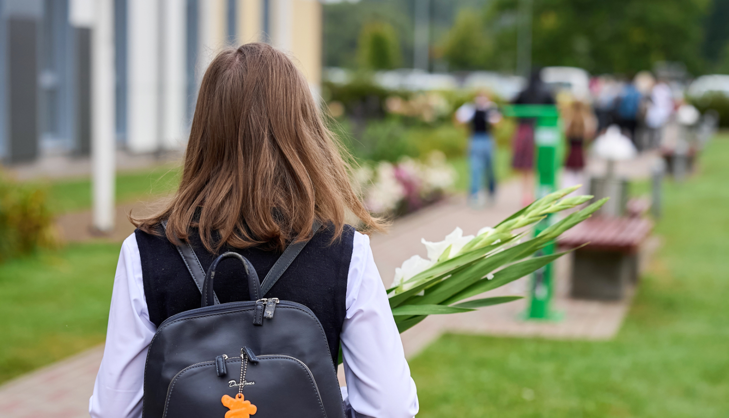 Skolniece ar melnu mugursomu no mugurpuses, rokās baltas gladiolas