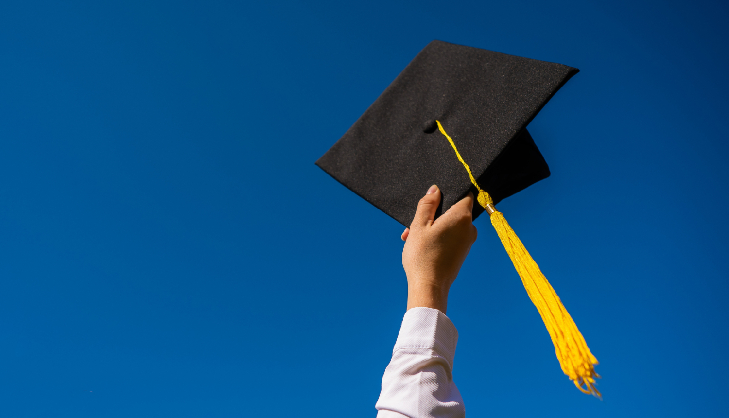 Rokā pacelta studenta izlaiduma cepure ar dzeltenu pušķi uz zila fona